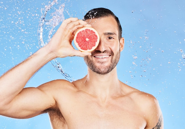 Portrait d'éclaboussures d'eau et de soins de la peau avec un beau jeune homme posant pour des soins corporels naturels Eau de douche et pamplemousse avec un modèle homme en studio sur fond bleu pour l'hygiène ou l'hydratation