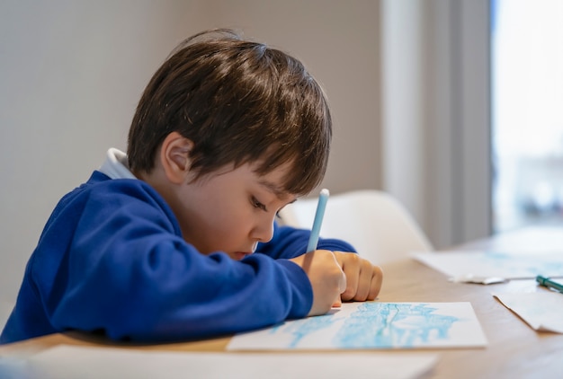 Portrait d'écolier assis seul à faire ses devoirs, enfant garçon tenant un stylo de couleur dessin et écriture sur papier blanc sur table, école élémentaire et concept homeschooling