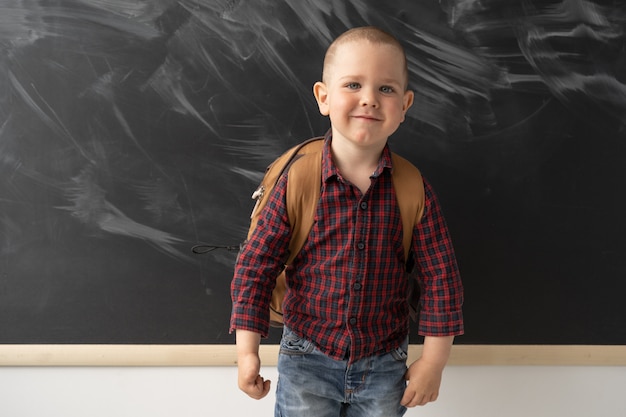 Portrait d'un écolier debout devant le tableau noir et souriant. Le tableau noir est propre avec des traces de craie. L'enfant est vêtu d'une chemise sombre et d'un jean. .