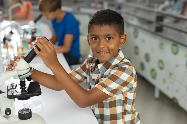 Photo portrait d'un écolier élémentaire biracial tenant un microscope alors qu'il est assis à son bureau dans un laboratoire