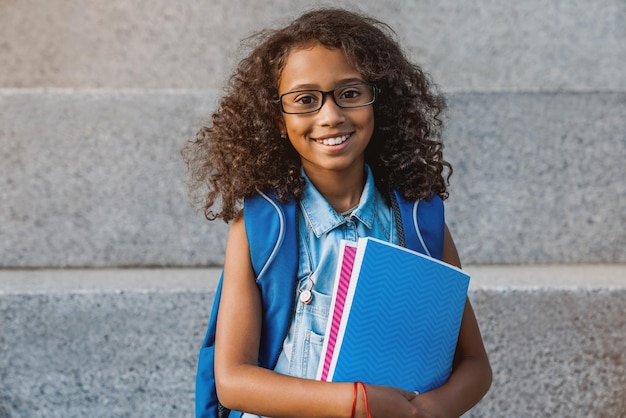 Portrait d'une écolière africaine heureuse avec sac à dos tenant des cahiers à l'extérieur de l'école primaire