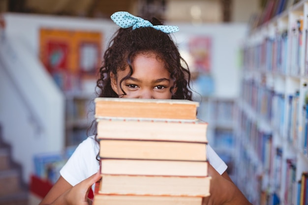 Portrait d'une écolière afro-américaine heureuse portant une pile de livres dans la bibliothèque de l'école