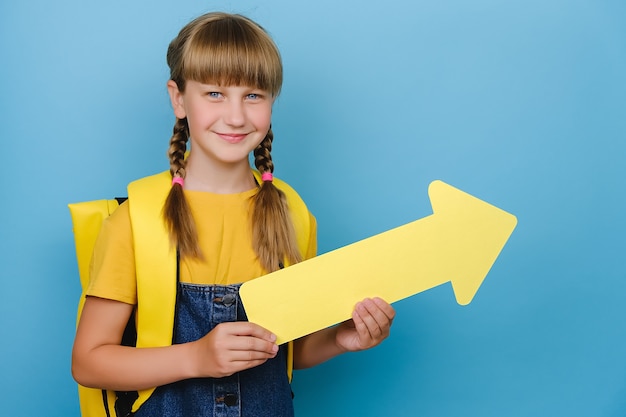 Portrait d'une écolière blonde souriante montrant de côté avec une flèche jaune, porte un sac à dos, posant isolé sur un mur de fond de couleur bleue avec un espace de copie pour le contenu promotionnel. Retour au concept de l'école