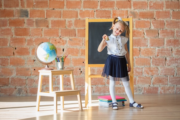 Portrait d'une écolière caucasienne ou d'une fille de la maternelle souriante devant le tableau noir avec une cloche dans les mains, concept de retour à l'école