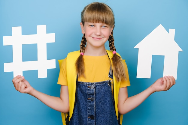 Portrait d'une écolière heureuse tenant un signe de hashtag blanc et un modèle de maison, étiquette pour les entreprises, porte un sac à dos jaune, posant sur fond bleu. Veille réseaux sociaux, mesure média, école