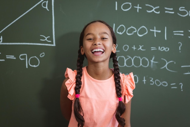 Portrait d'une écolière métisse en train de rire debout devant un tableau dans une leçon de mathématiques en classe