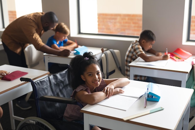 Portrait d'une écolière primaire biraciale souriante assise sur un fauteuil roulant à un bureau en classe.