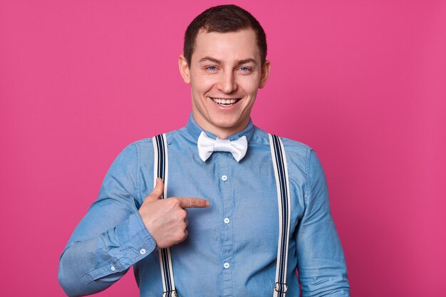 Portrait de l'élégant beau jeune homme posant isolé sur mur rose. Beau mâle souriant et pointant de côté avec son index, un gars portant une chemise bleue, des bretelles et un noeud papillon blanc.