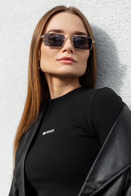 Portrait élégant d'une belle jeune femme caucasienne modèle avec des lunettes dorées vintage à la mode dans des vêtements de mode avec une veste en cuir noir et un t-shirt sur un mur de fond blanc dans la ville