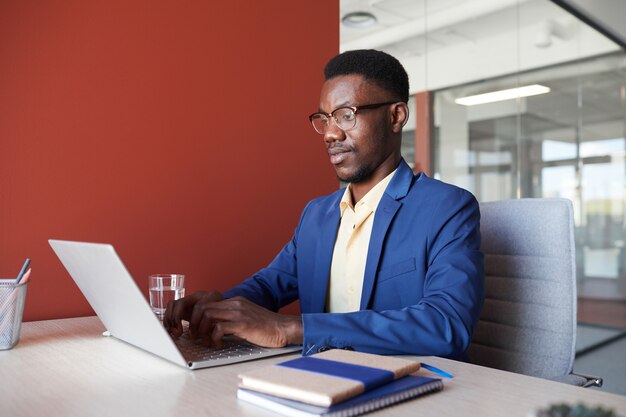 Portrait de l'élégant homme afro-américain utilisant un ordinateur portable au bureau tout en travaillant dans l'intérieur de bureau contemporain, copiez l'espace