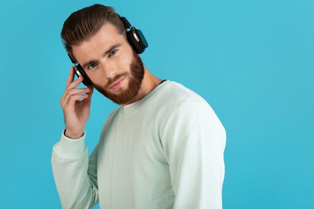 Portrait de l'élégant jeune homme barbu attrayant, écouter de la musique sur des écouteurs sans fil style moderne humeur confiante isolé sur fond bleu
