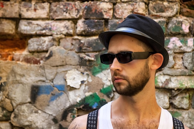 Portrait élégant jeune homme barbu à lunettes de soleil avec bretelles et chapeau au fond de mur de brique