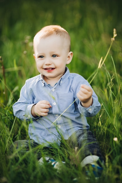 Portrait de l'élégant petit garçon blond assis dans le parc parmi l'herbe