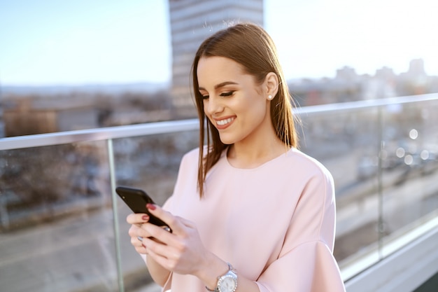 Portrait de l'élégante belle brune avec un sourire à pleines dents à l'aide d'un téléphone intelligent sur la terrasse