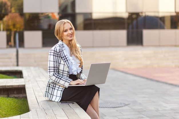 Portrait d'élégante belle femme d'affaires confiante aux cheveux blonds en veste à carreaux et jupe assis sur un banc et en tapant sur un ordinateur portable à l'air positif satisfait à la caméra travaillant en freelance en plein air