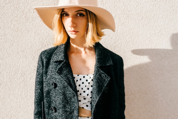 Portrait d'élégante élégante fille blonde cheveux courts en chapeau et veste posant sur fond beige