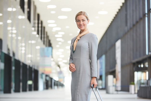 Portrait d'une élégante hôtesse de l'air blonde avec valise et souriant tout en posant à l'aéroport,