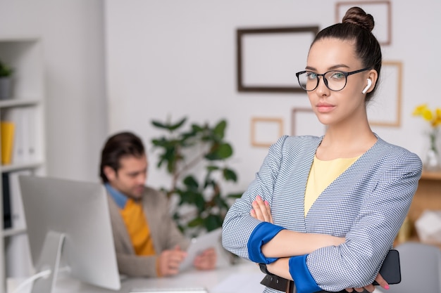 Portrait de l'élégante jeune femme d'affaires dans les écouteurs sans fil debout avec les bras croisés dans un bureau moderne, homme travaillant en arrière-plan