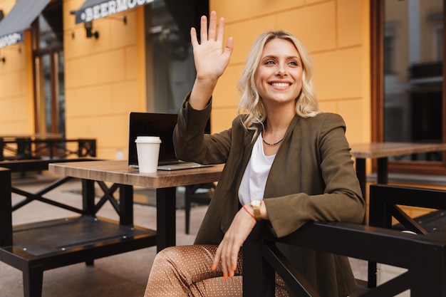 Portrait d'élégante jeune femme en agitant la main de côté, tout en travaillant sur un ordinateur portable dans un café en plein air