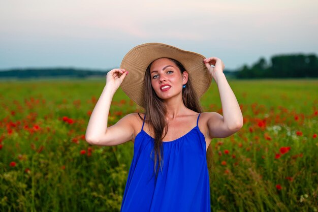 Portrait d'une élégante jeune femme aux cheveux longs et chapeau de paille dans un champ de coquelicots au soleil du soir