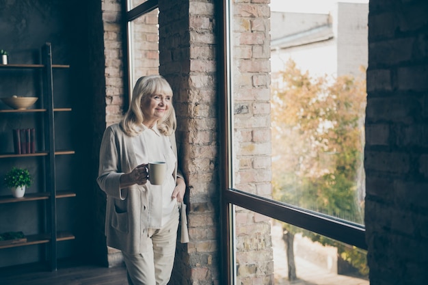 Portrait d'elle elle belle belle attrayante belle blonde aux cheveux gris rêveuse mamie d'âge moyen regardant la fenêtre au repos boire du cacao au loft industriel en brique de style moderne intérieur maison appartement