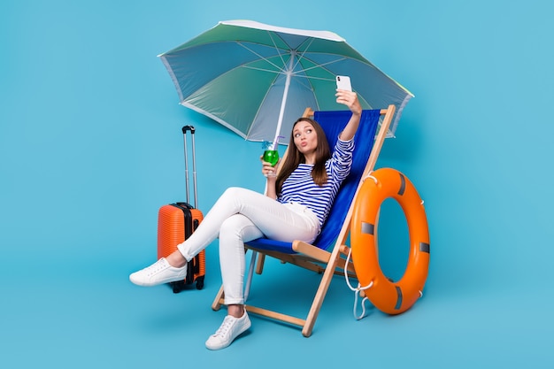 Portrait d'elle elle belle jolie fille gaie et funky assise sur une chaise sous un parasol buvant un mojito exotique faisant un selfie envoyant un baiser d'air isolé sur fond de couleur bleu vif brillant éclatant