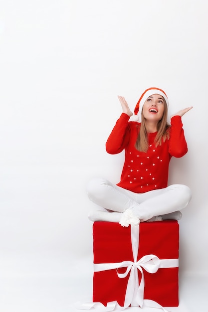 Portrait émotionnel d'une fille dans un chapeau de Noël assis sur une boîte-cadeau rouge avec un arc blanc