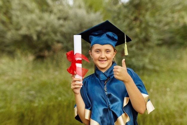 Photo portrait émotionnel d'une petite étudiante diplômée heureuse dans une robe de graduation bleue avec chapeau, titulaire d'un diplôme, en plein air. écolière gaie chanceuse célébrant le triomphe.