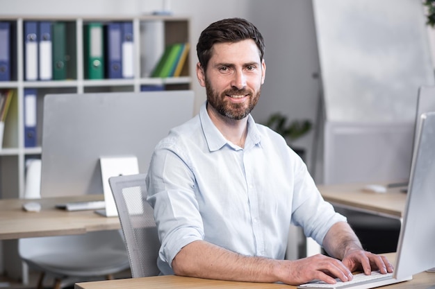 Portrait d'un employé de bureau Il regarde la caméra sourit Assis à un bureau au bureau à l'ordinateur dans une chemise blanche