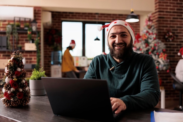 Portrait d'un employé de bureau travaillant sur un ordinateur portable de l'entreprise et assis dans un bureau décoré d'arbres de noël et de lumières, d'ornements festifs. Homme souriant portant un bonnet de Noel pendant la saison de Noël.