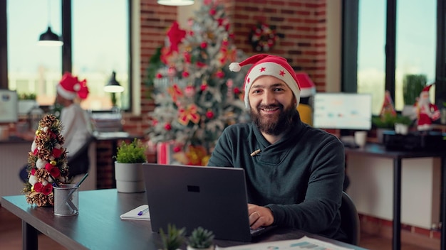 Portrait d'un employé de bureau utilisant un ordinateur portable au bureau pendant la saison des vacances d'hiver, portant un bonnet de noel au travail. Employé de sexe masculin travaillant sur ordinateur avant de célébrer la fête de la veille de noël.