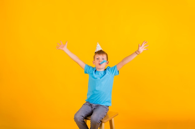 Portrait d'un enfant en bas âge garçon assis dans un chapeau de vacances en papier et sifflet et étend ses mains sur les côtés sur un fond jaune