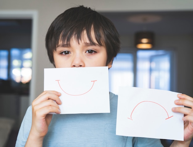 Portrait d'un enfant ennuyé avec un visage triste tenant du papier blanc avec le sourire et la tristesse, un enfant garçon s'ennuyant reste à la maison pendant l'auto-isolement, la quarantaine. Épidémie de coronavirus et épidémie de grippe