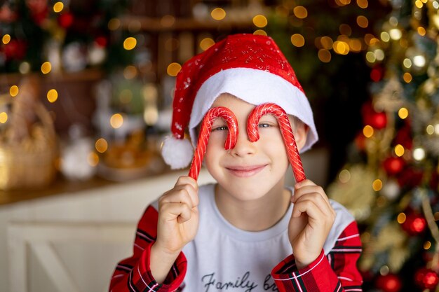 Portrait d'un enfant garçon heureux dans un chapeau de Père Noël dans la cuisine avec un décor de Noël rouge avec de gros bonbons et souriant en prévision des vacances