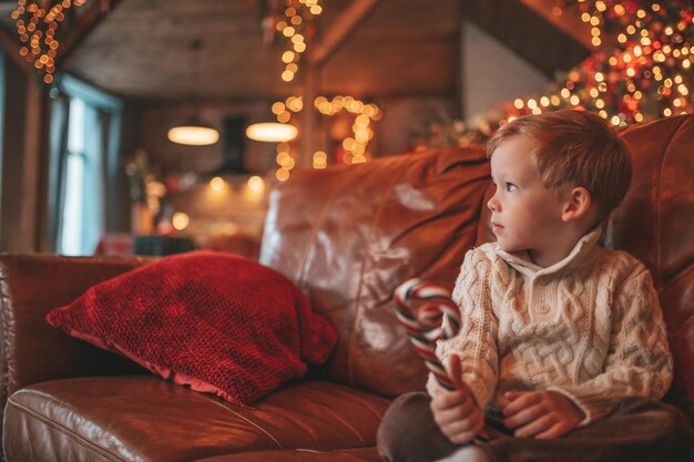 Portrait d'un enfant heureux candide en pull beige en tricot tenir une tasse de Noël avec des guimauves et une canne en bonbon