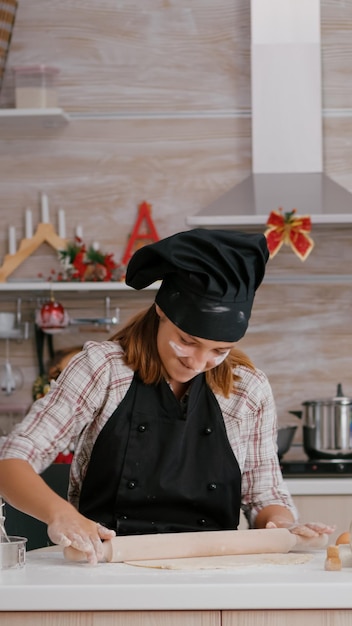 Portrait d'un enfant heureux portant un tablier faisant de la pâte maison à l'aide d'un rouleau à pâtisserie préparant un délicieux dessert traditionnel au pain d'épice. Fille debout à table dans une cuisine décorée de Noël profitant de Noël