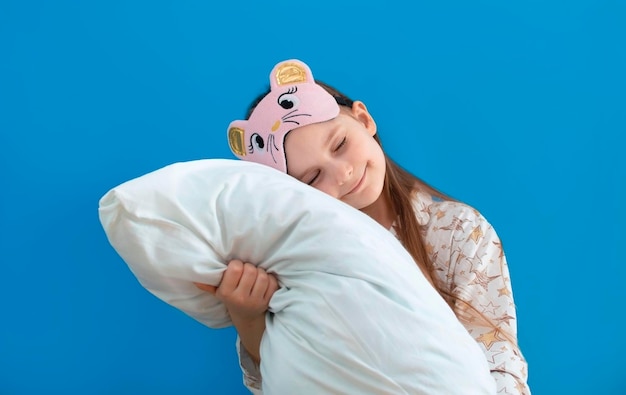 Photo portrait d'un enfant joyeux positif dans un masque pour les yeux étreignant son gros oreiller doux sur fond bleu profitant de temps libre le week-end se sentant satisfait d'aller au lit porter un pyjama blanc