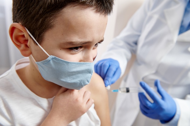 Portrait d'un enfant de sexe masculin effrayé dans un masque médical recevant une dose de vaccin contre les maladies infectieuses