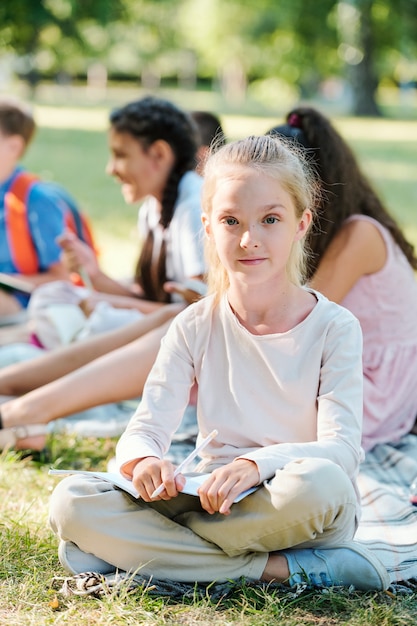 Portrait d'un enseignant souriant écoutant une petite fille faisant une présentation assise avec un groupe d'enfants et profitant d'une leçon en plein air