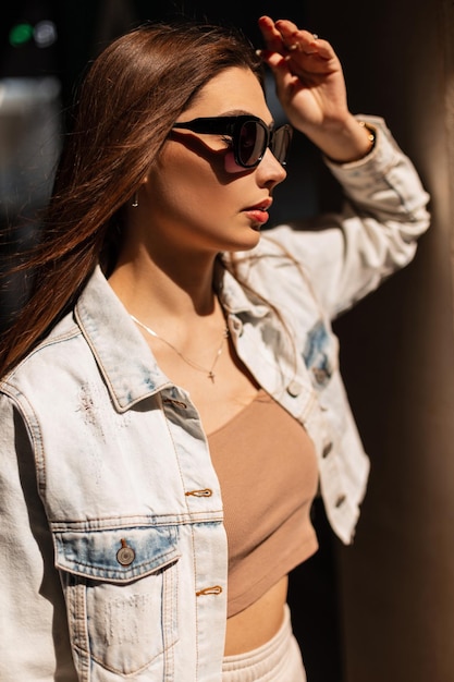 Portrait ensoleillé d'un joli modèle féminin avec des lunettes de soleil de mode en veste de jeans et haut se dresse dans la rue au soleil