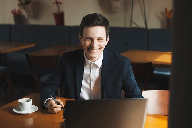 Portrait d'un entrepreneur caucasien confiant positif travaillant à son ordinateur portable tout en souriant assis dans un café.