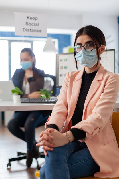 Portrait d'un entrepreneur gestionnaire avec un masque facial comme mesure de sécurité pendant l'épidémie de coronavirus en gardant une distance sociale avec les collègues dans un immeuble de bureaux en regardant la caméra.