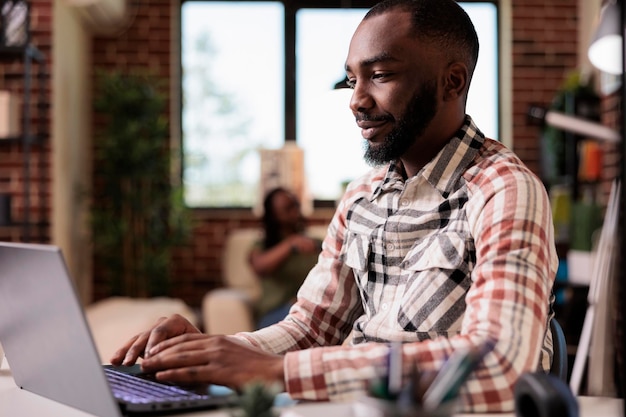 Portrait d'un entrepreneur travaillant à distance en tapant et en regardant l'écran d'un ordinateur portable pendant que sa femme regarde la télévision depuis un canapé. Pigiste afro-américain utilisant un ordinateur portable pendant que le colocataire se détend.