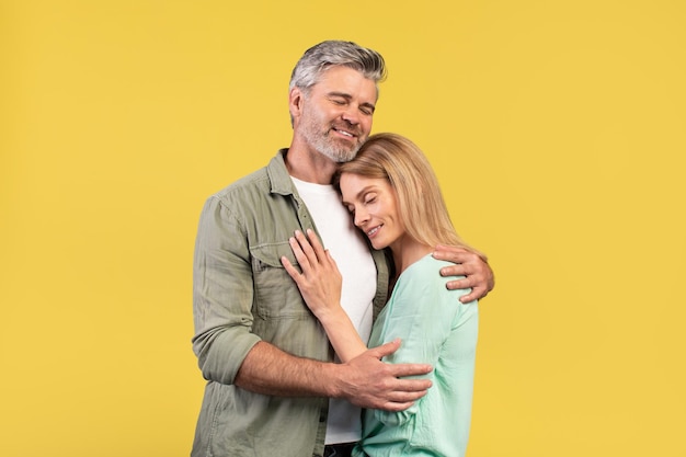 Portrait d'époux d'âge moyen aimants serrant les yeux fermés tout en se tenant isolés sur fond de studio jaune