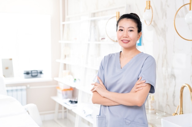 Portrait de l'esthéticienne asiatique d'âge moyen positif en gommages debout avec les bras croisés dans la salle de soins de beauté avec un design contemporain