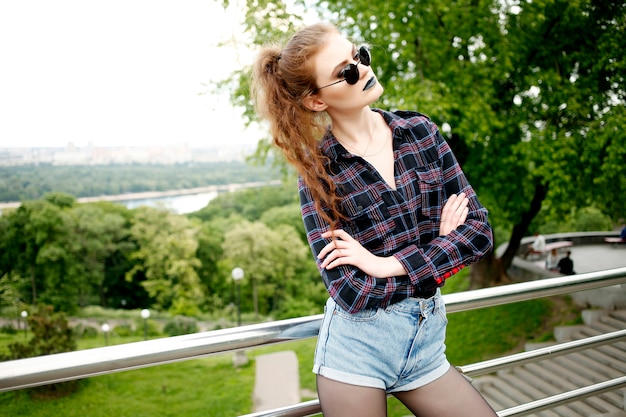 Portrait d'été de belle fille hipster maigre dans une chemise à carreaux et un short en jeans dans des lunettes de soleil élégantes et du rouge à lèvres bleu