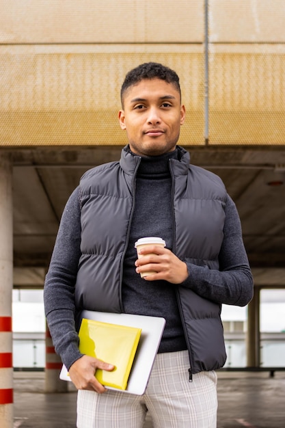 Portrait d'un étudiant universitaire latin souriant avec un ordinateur portable à café et des documents à l'extérieur