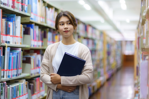 Portrait d'étudiant à l'université de femme asiatique intelligente livre de lecture et regardant la caméra entre les étagères dans la bibliothèque du campus avec copyspace.