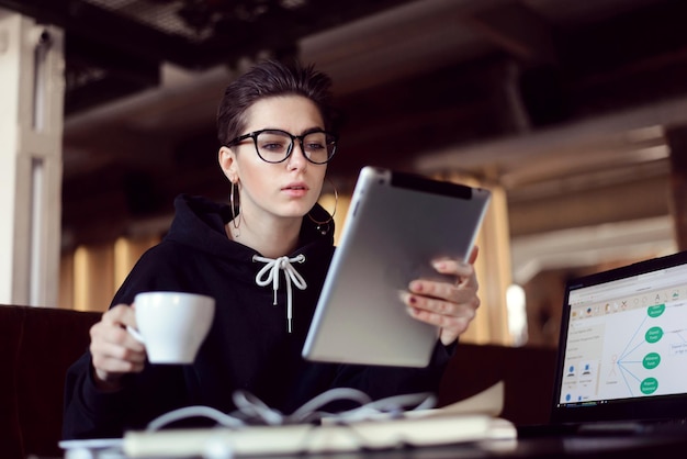 Portrait d'une étudiante sérieuse aux cheveux courts portant des lunettes assise avec la tablette pendant la pause au café