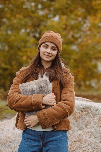 Portrait d'une étudiante souriante de l'enseignement supérieur avec un ordinateur portable debout dans le parc à l'automne